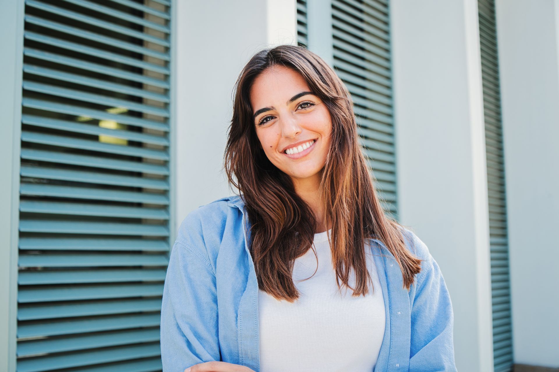 Woman smiling, standing outdoors. Wearing a light blue jacket over a white top. Behind her are gray horizontal shutters.
