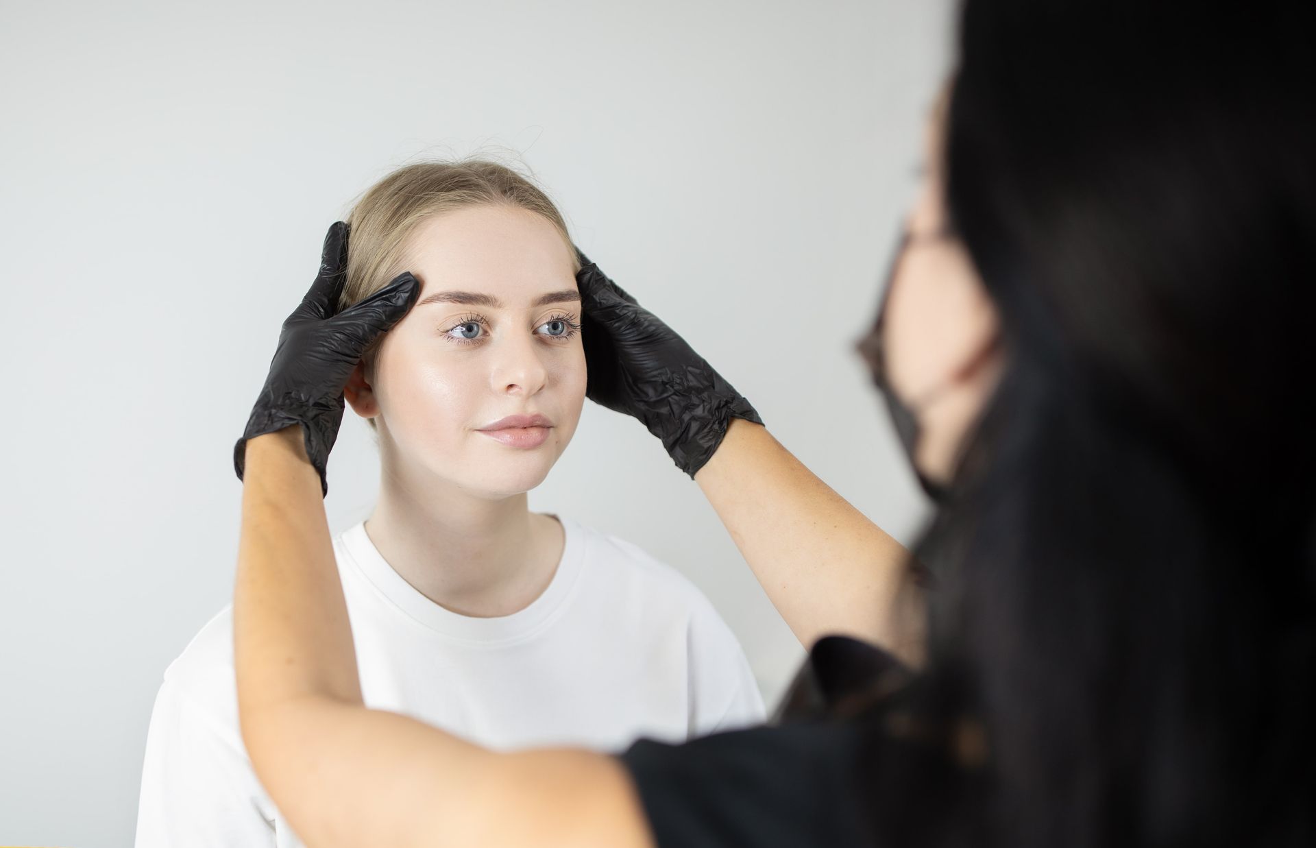 Woman getting face examined by a person wearing a mask and gloves in a white room.