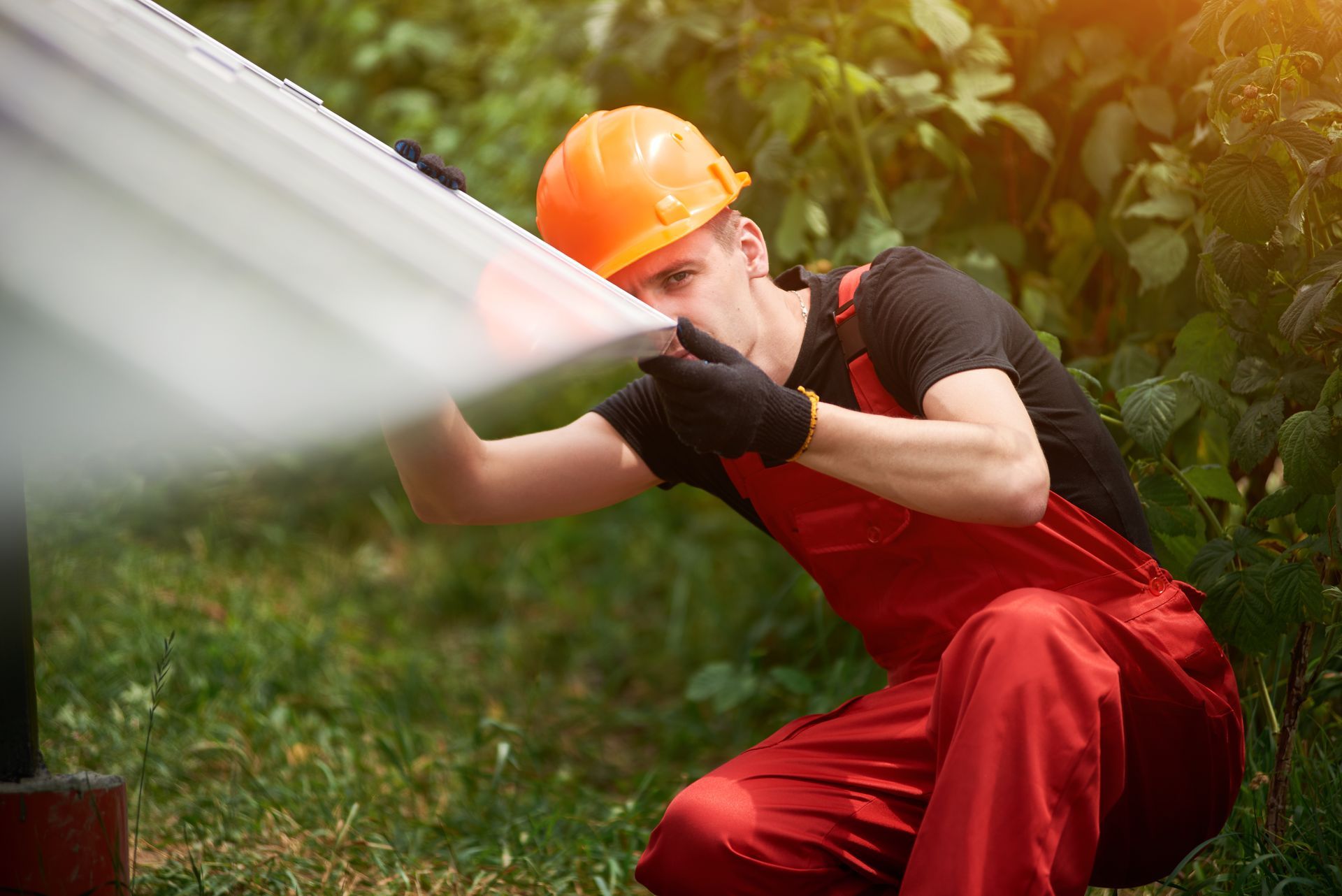 Man in orange hard hat and red overalls installing a solar panel outdoors.