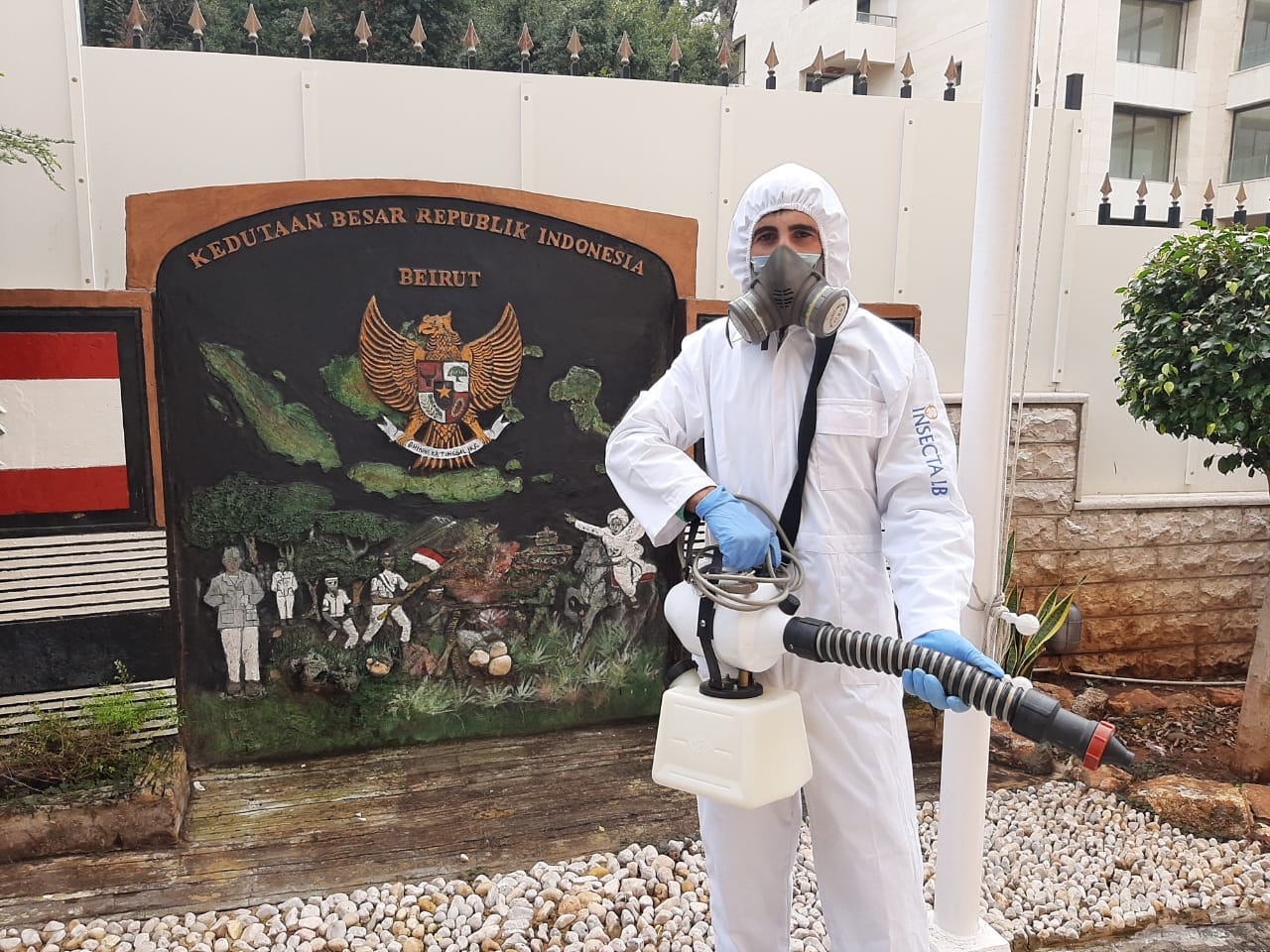 Person in protective suit spraying disinfectant near a monument with a relief of the Indonesian coat of arms.