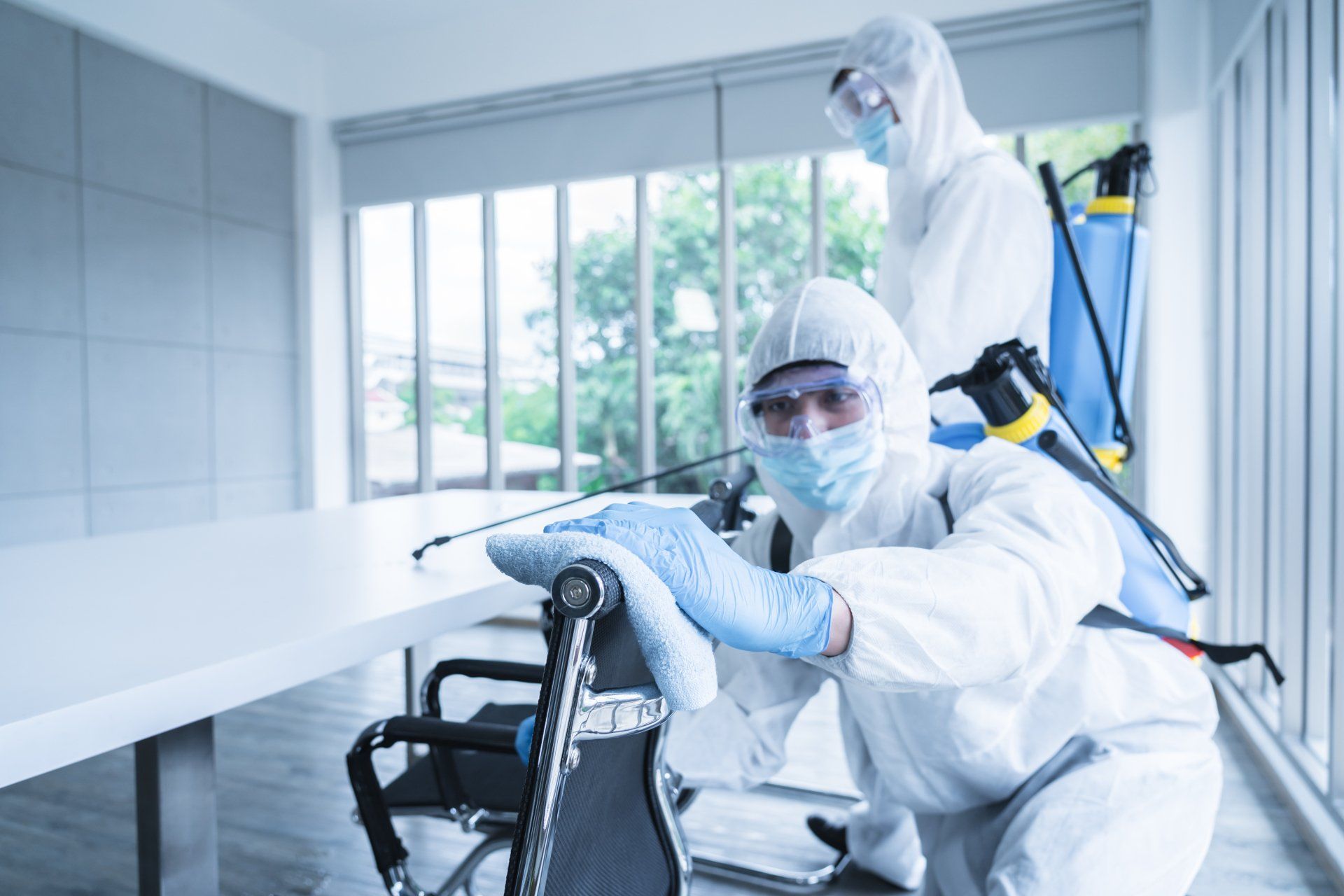 Two people in protective suits sanitizing an office with sprayers; one person is kneeling and spraying.
