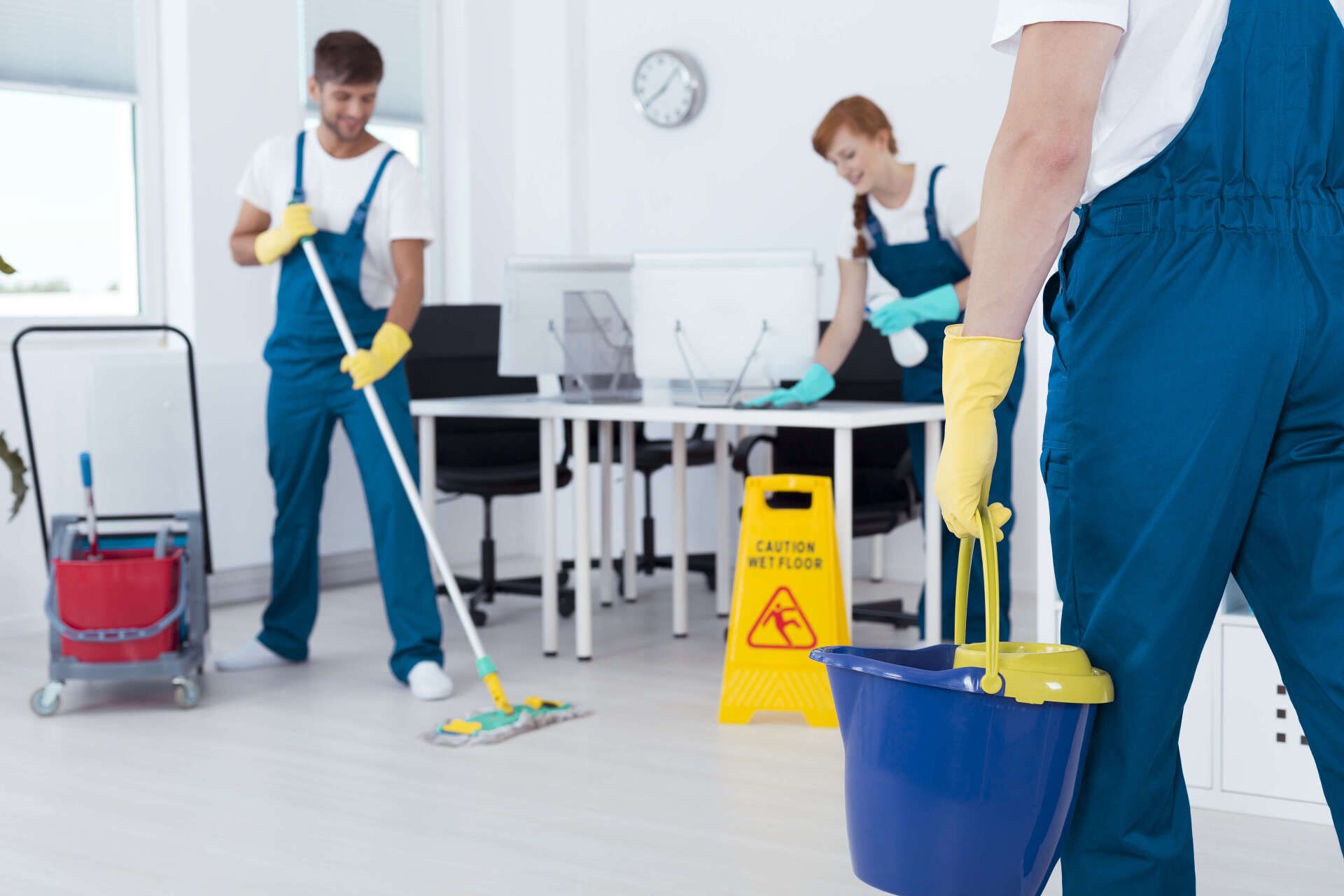 Three cleaning staff in blue overalls clean an office. One mops the floor, another dusts, and the third carries a bucket.