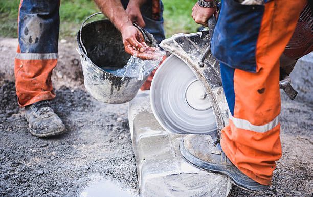 Workers cutting a stone block with a saw, using water. Blue and orange work pants, outdoor setting.