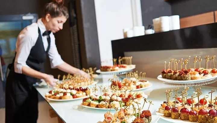 A server in a vest and white shirt arranges a buffet table filled with various colorful appetizers on skewers.