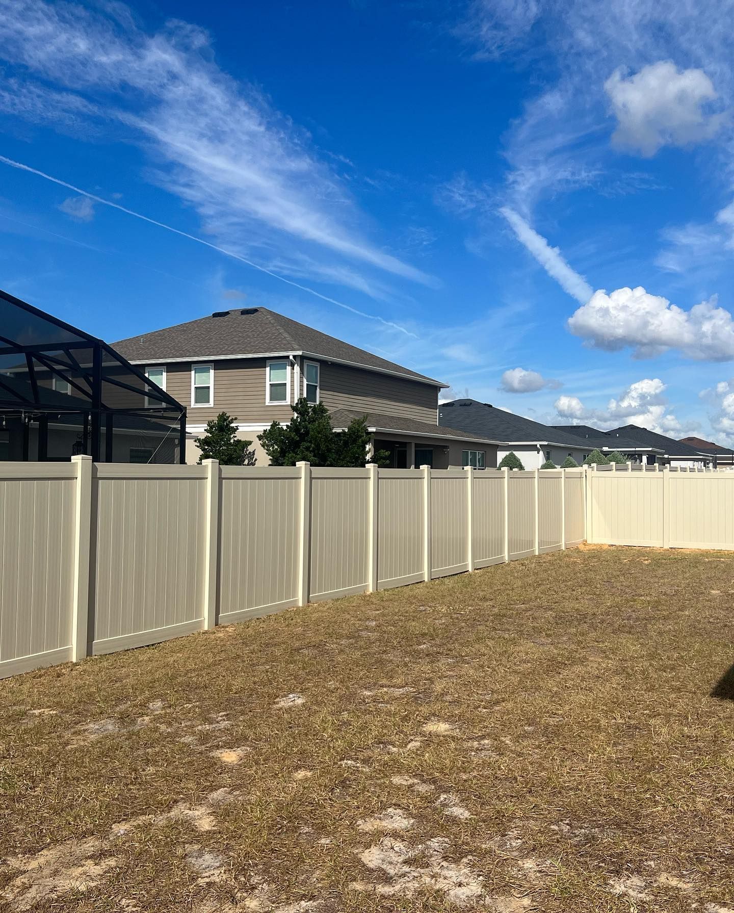Tan vinyl fence in a backyard, houses in background, blue sky with clouds.