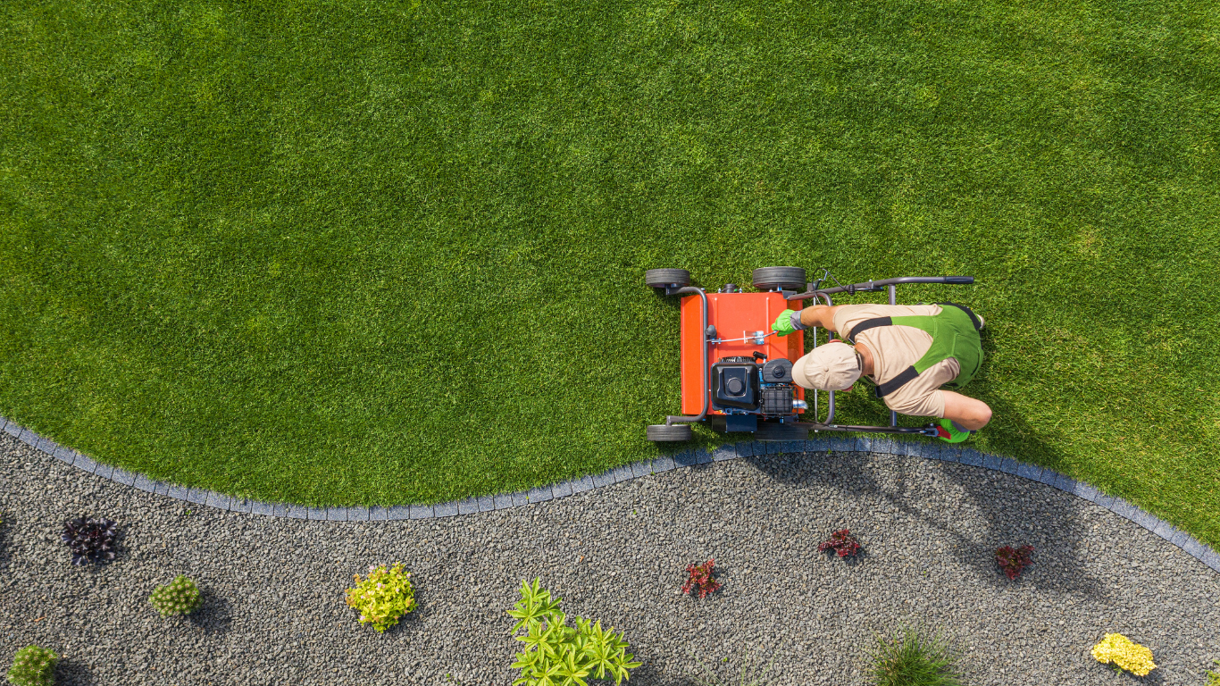 A person mowing a lush green lawn with a red mower next to a gravel border.