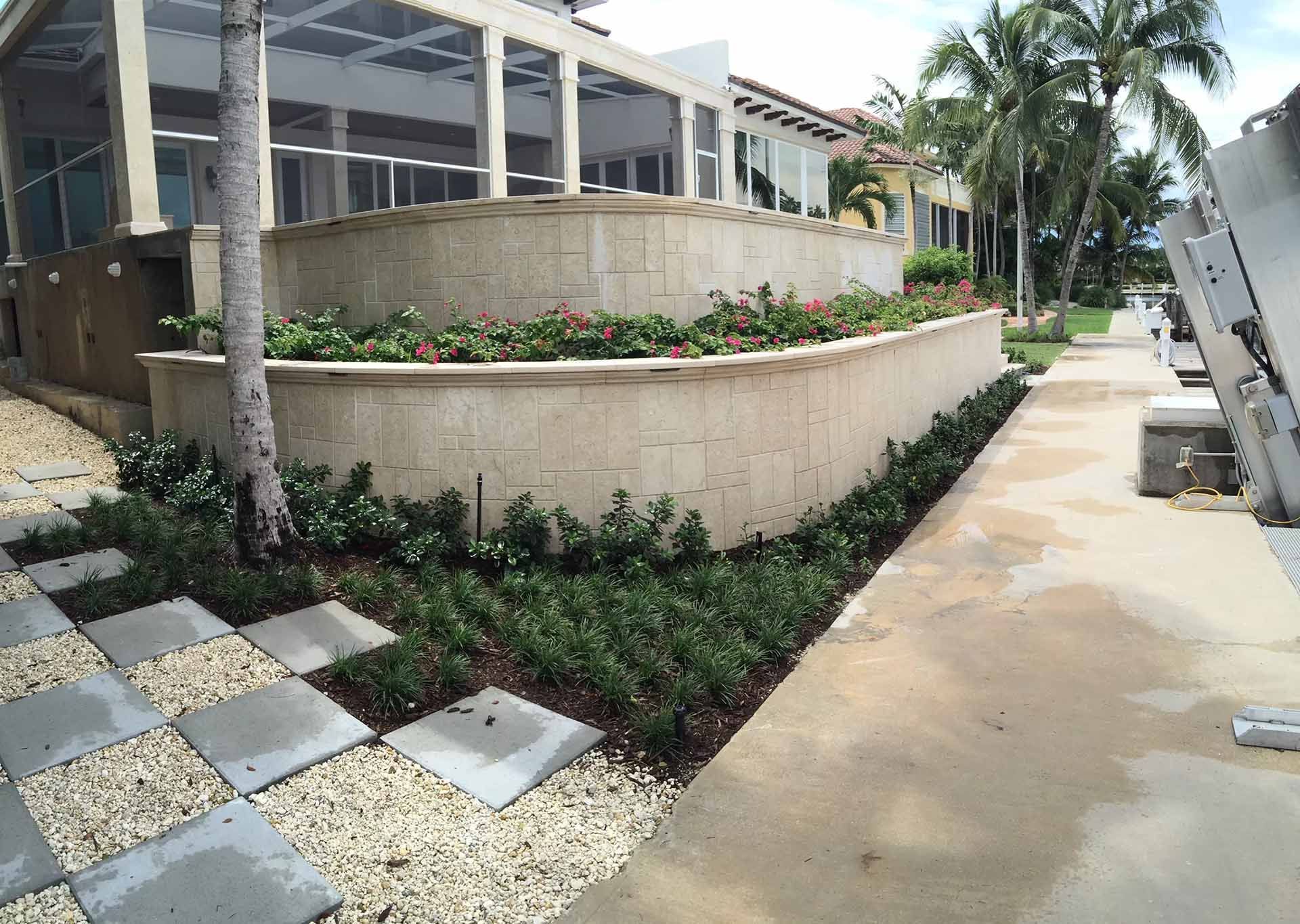 Exterior of a building with tiered beige stone planters with red flowers, a paved walkway, and landscaping