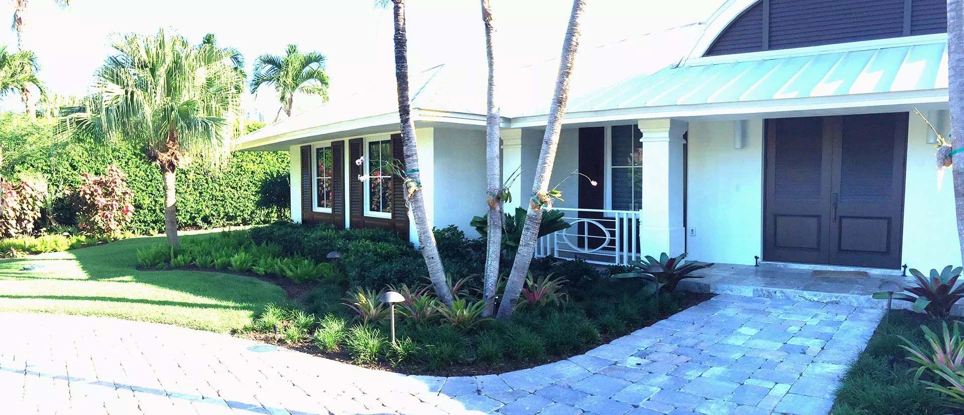 A white house with a green roof, a brick pathway, and palm trees