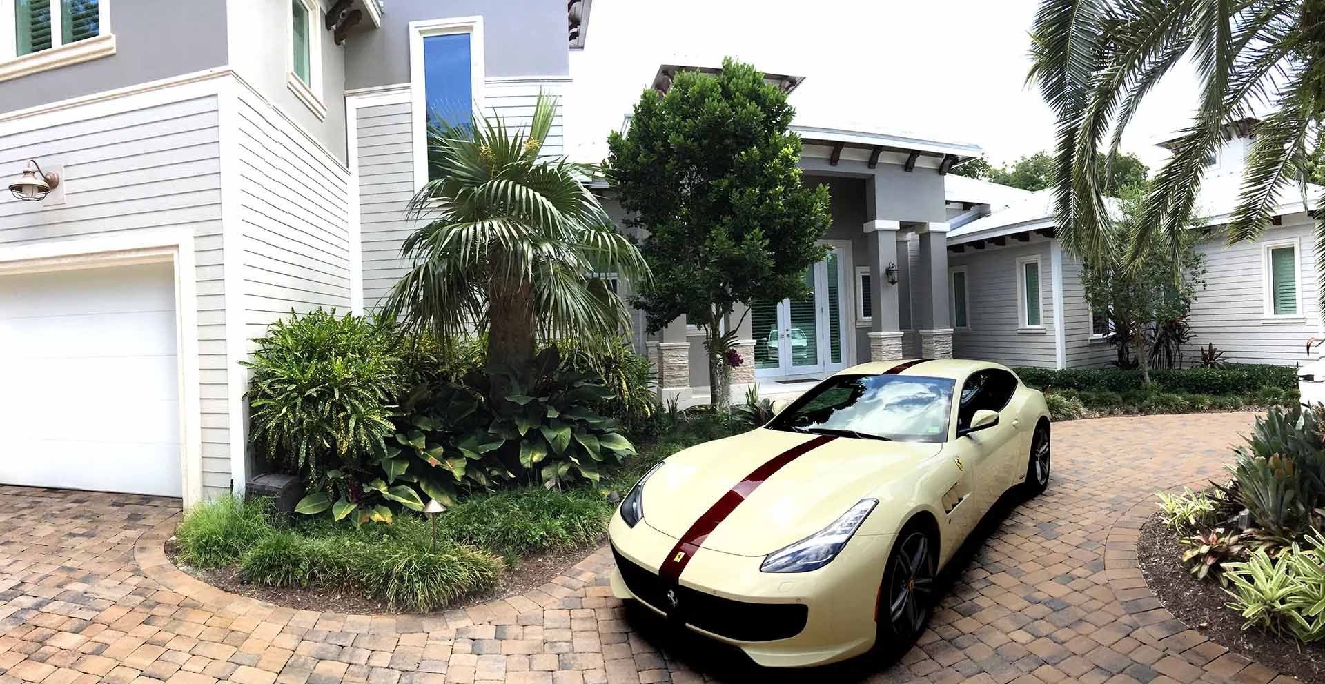 Tan sports car with dark racing stripe parked in front of a large house with a brick driveway