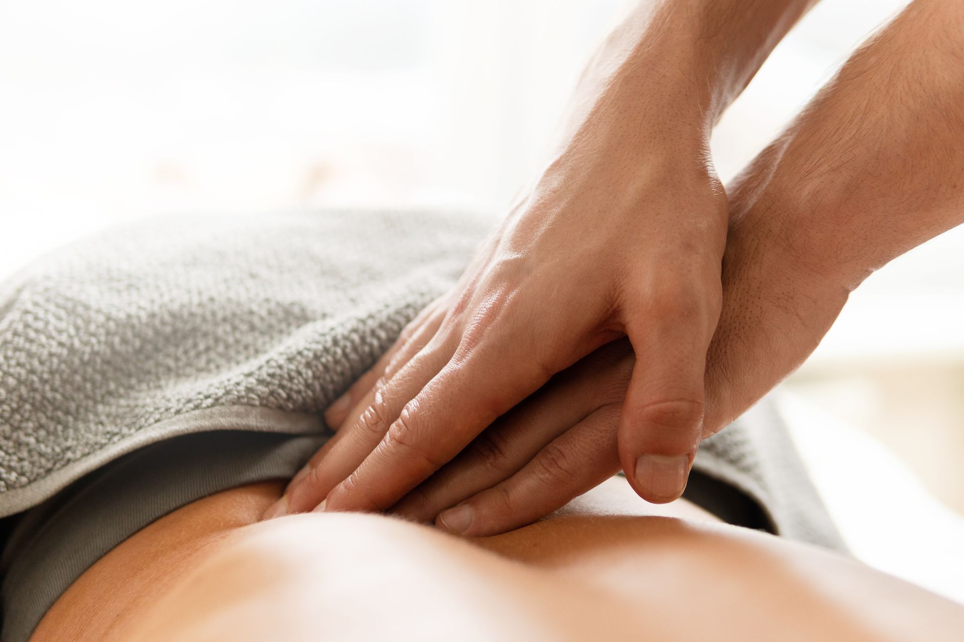 Hands massaging a person's lower back, covered by a gray towel, in a well-lit setting.