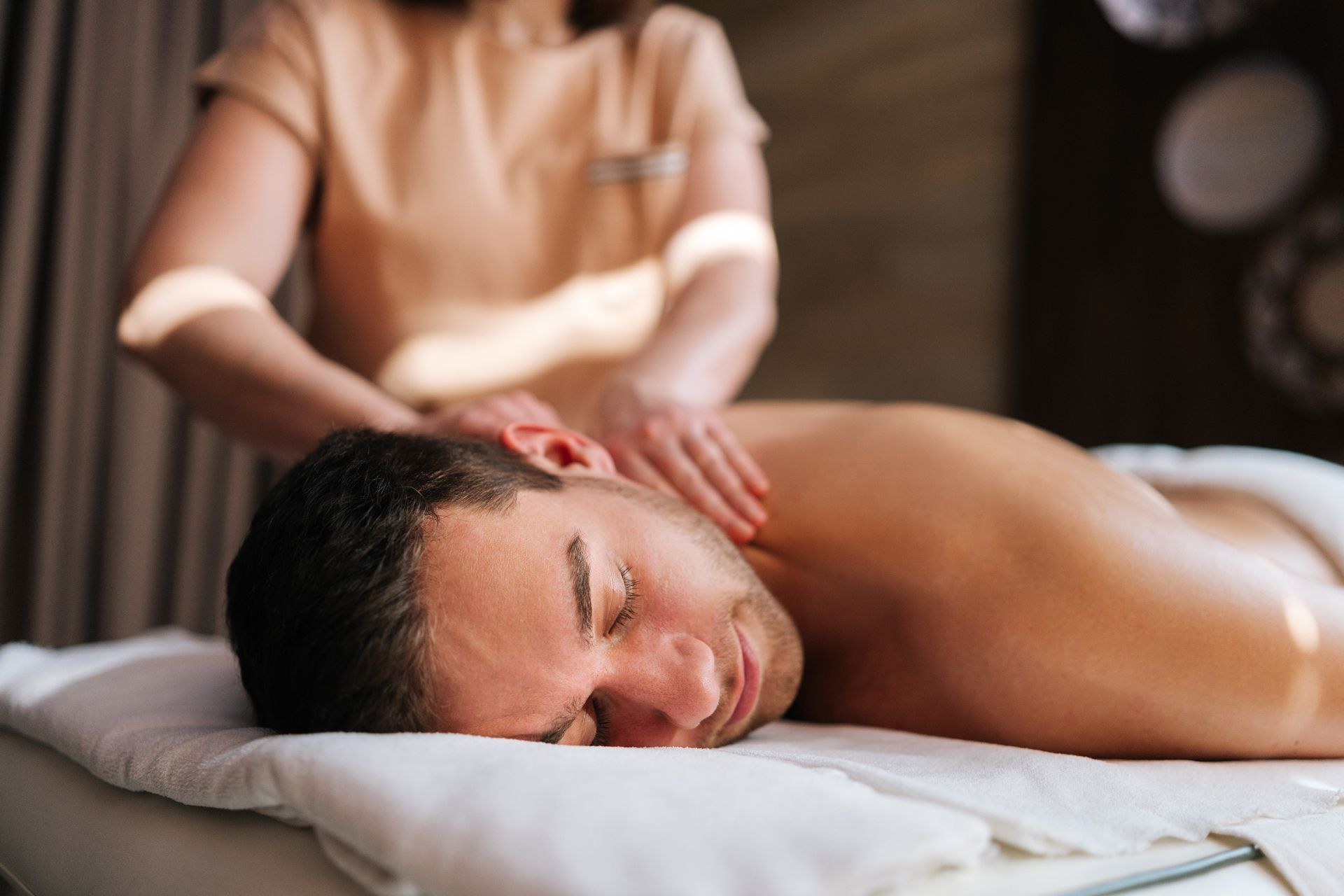 Man receiving shoulder massage at a spa; masseuse's hands on his neck.