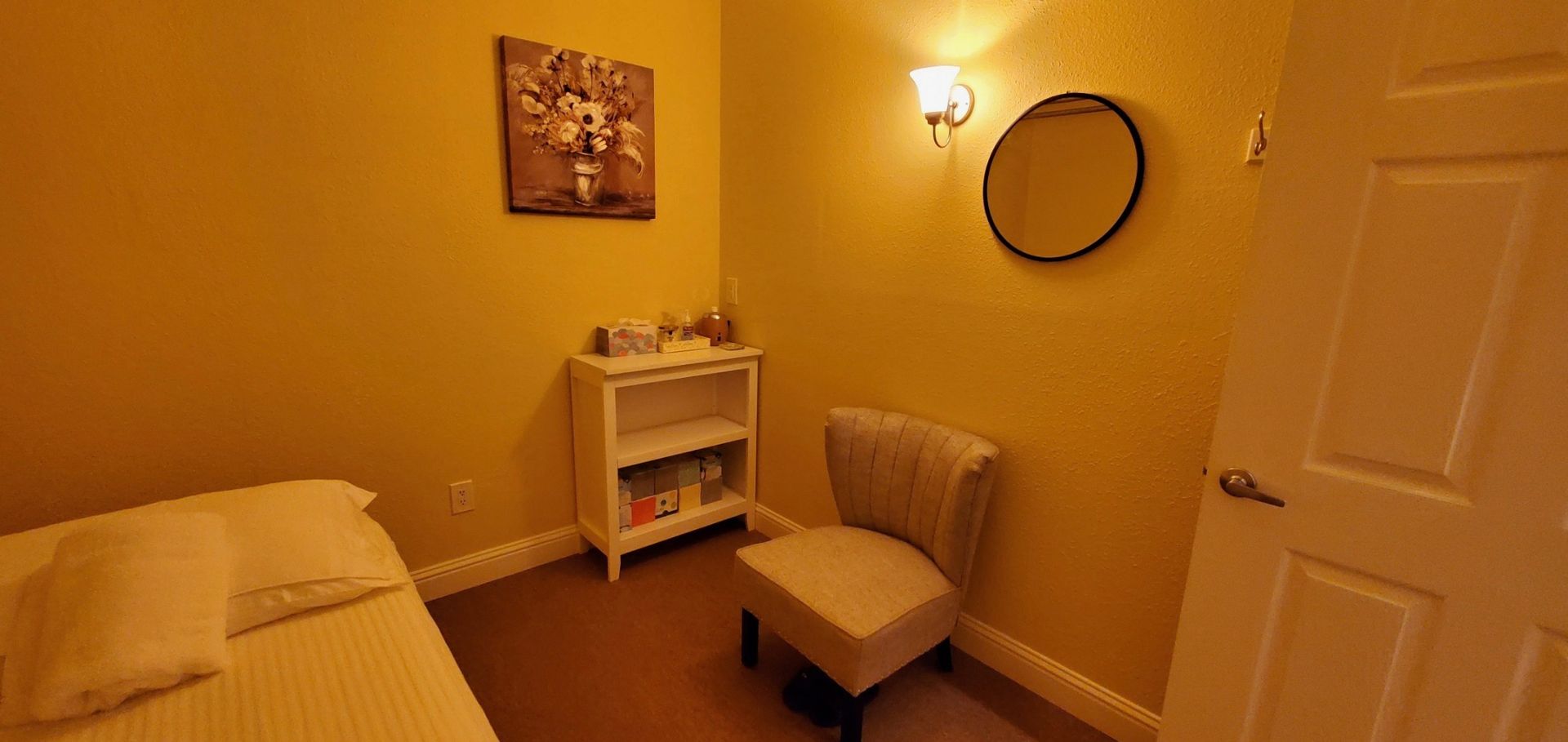 A massage room with a bed, chair, shelves, a round mirror, and a door. The walls are yellow.