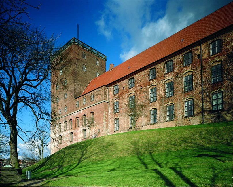 En stor murstensbygning med rødt tag ligger på toppen af ​​en græsklædt bakke