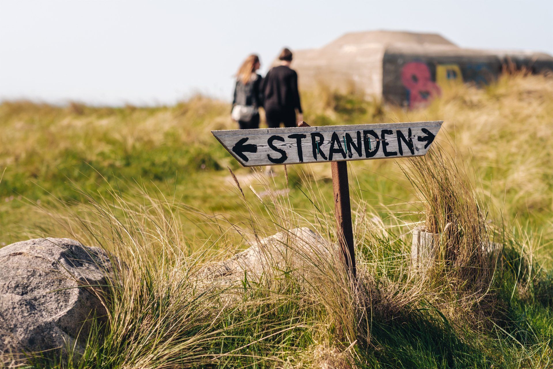 Et skilt i græsset peger mod stranden.