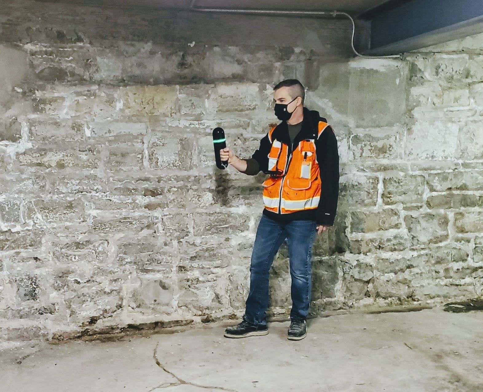 A man wearing a mask is standing in front of a stone wall holding a microphone.