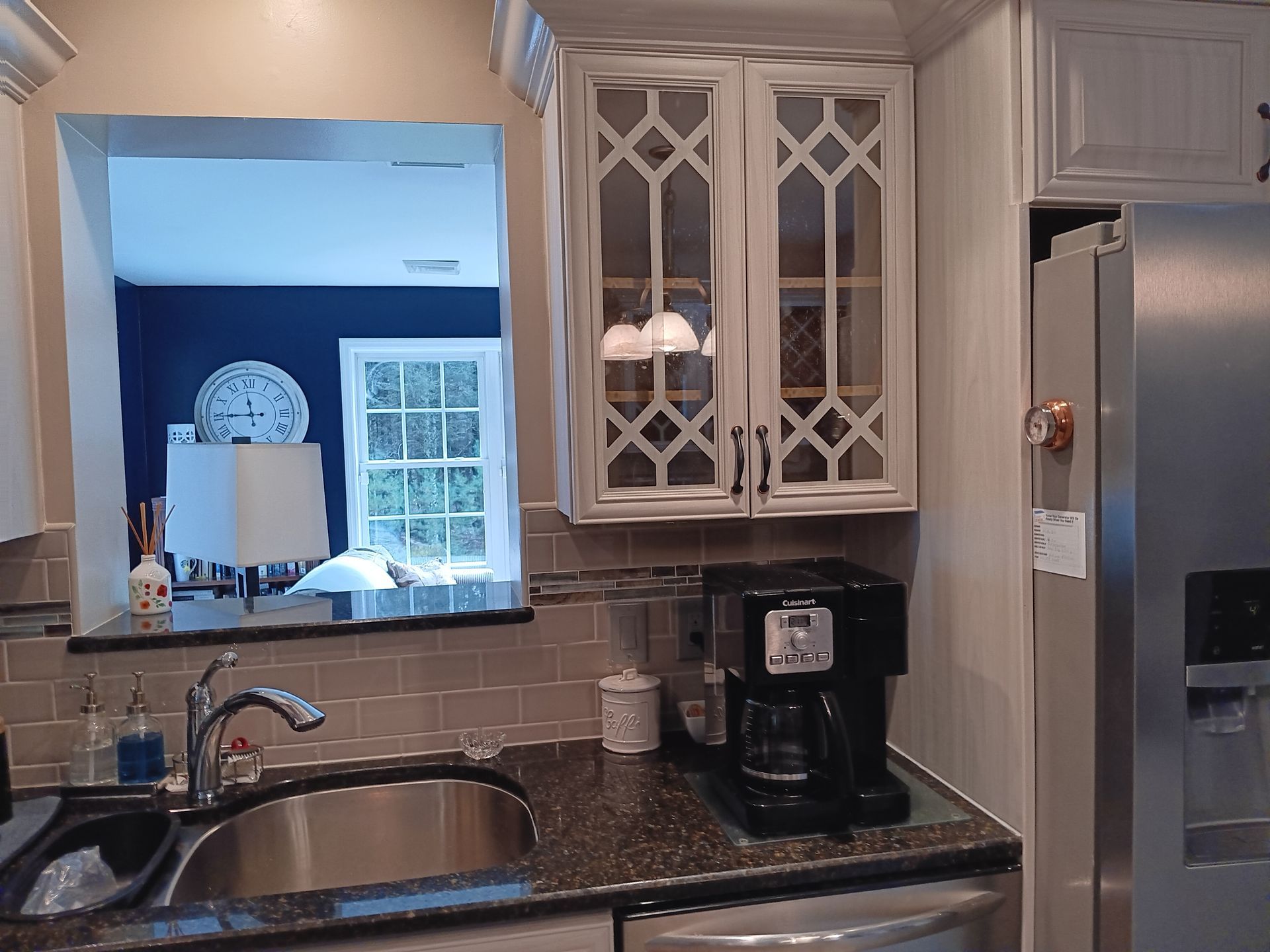 Kitchen with sink, coffee maker, and cabinets. A window reveals a room with a clock and lamp.