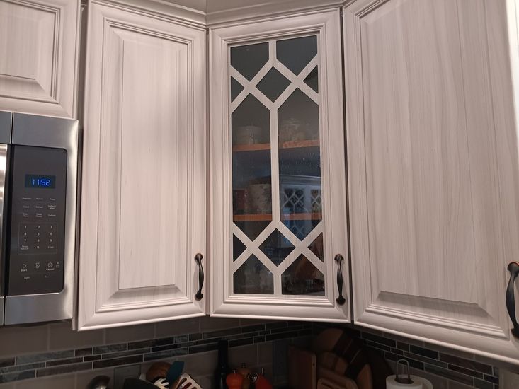 White kitchen cabinets with a glass-paned door, above a microwave and tiled backsplash.