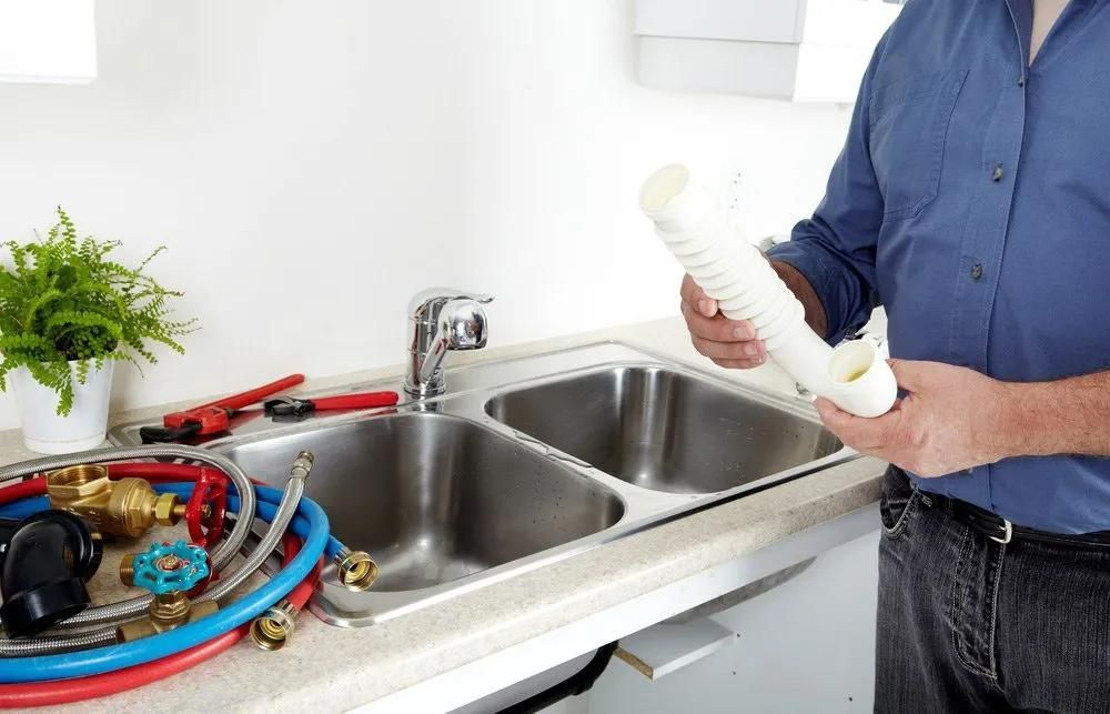 A Man Is Standing in Front of A Kitchen Sink Holding a Pipe — Gillespie Plumbing in Grafton, NSW