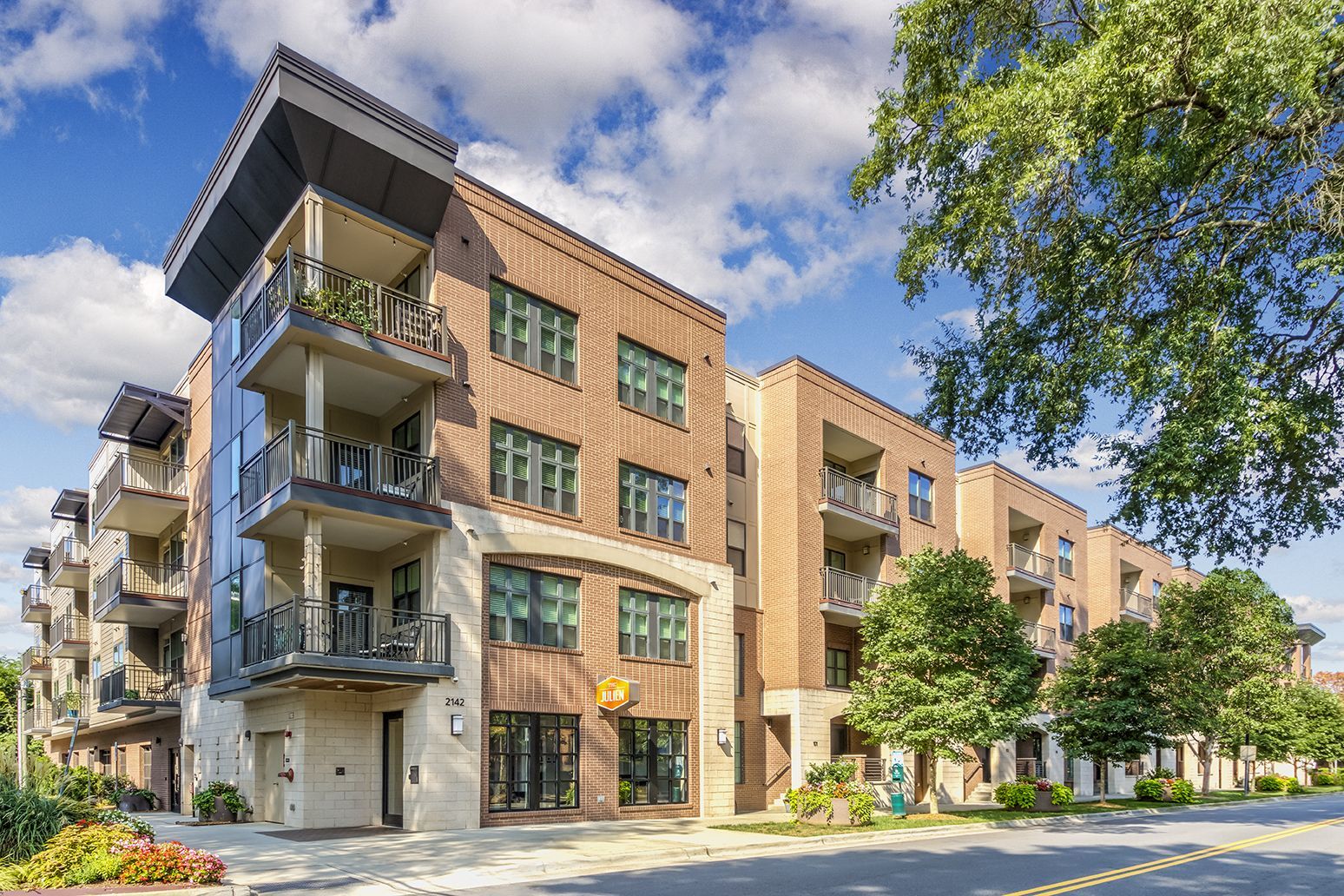 Multi-story brick apartment building with balconies, trees, and blue sky.