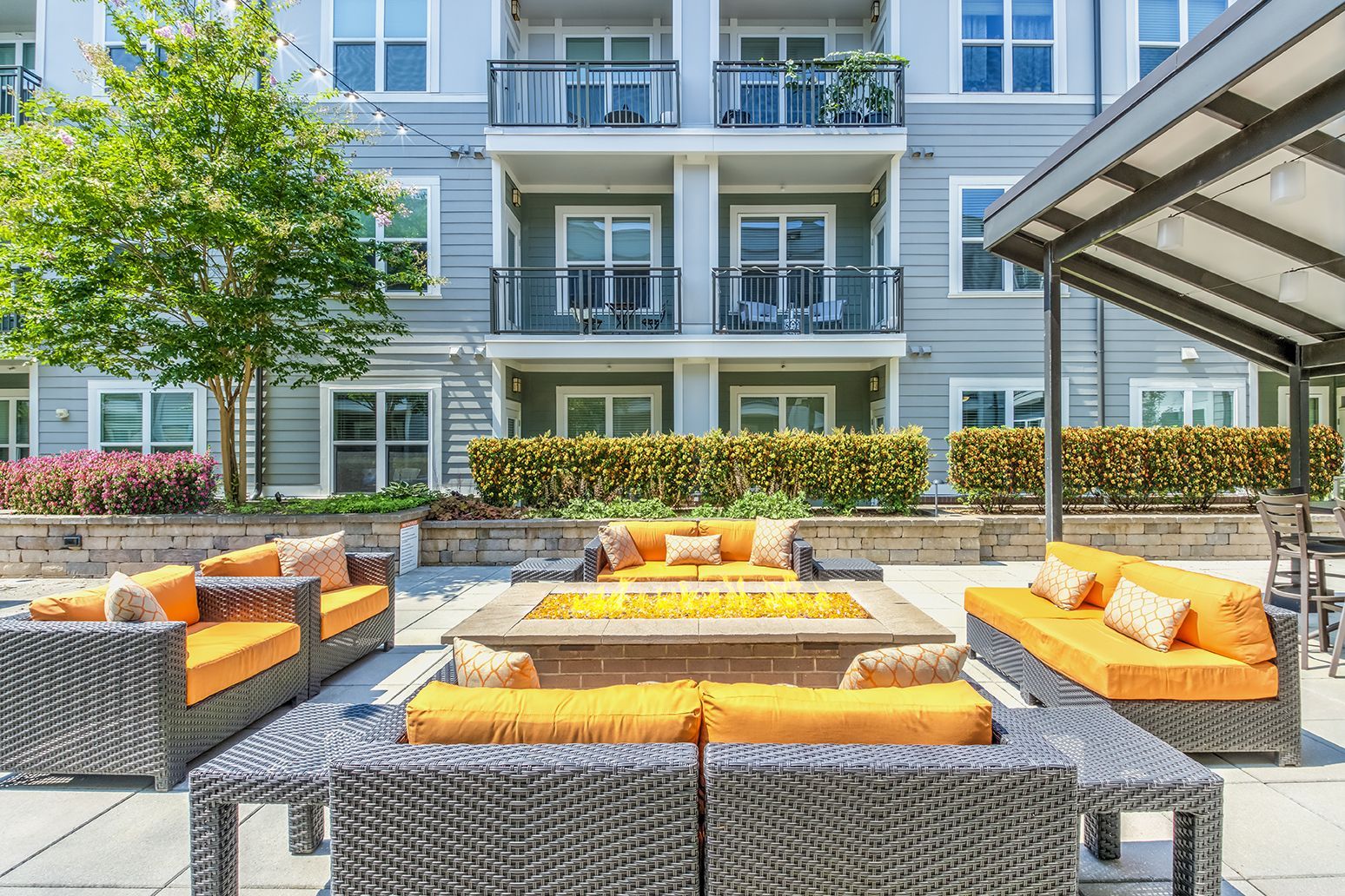 Outdoor seating area with fire pit and orange cushions, in front of a light blue apartment building.