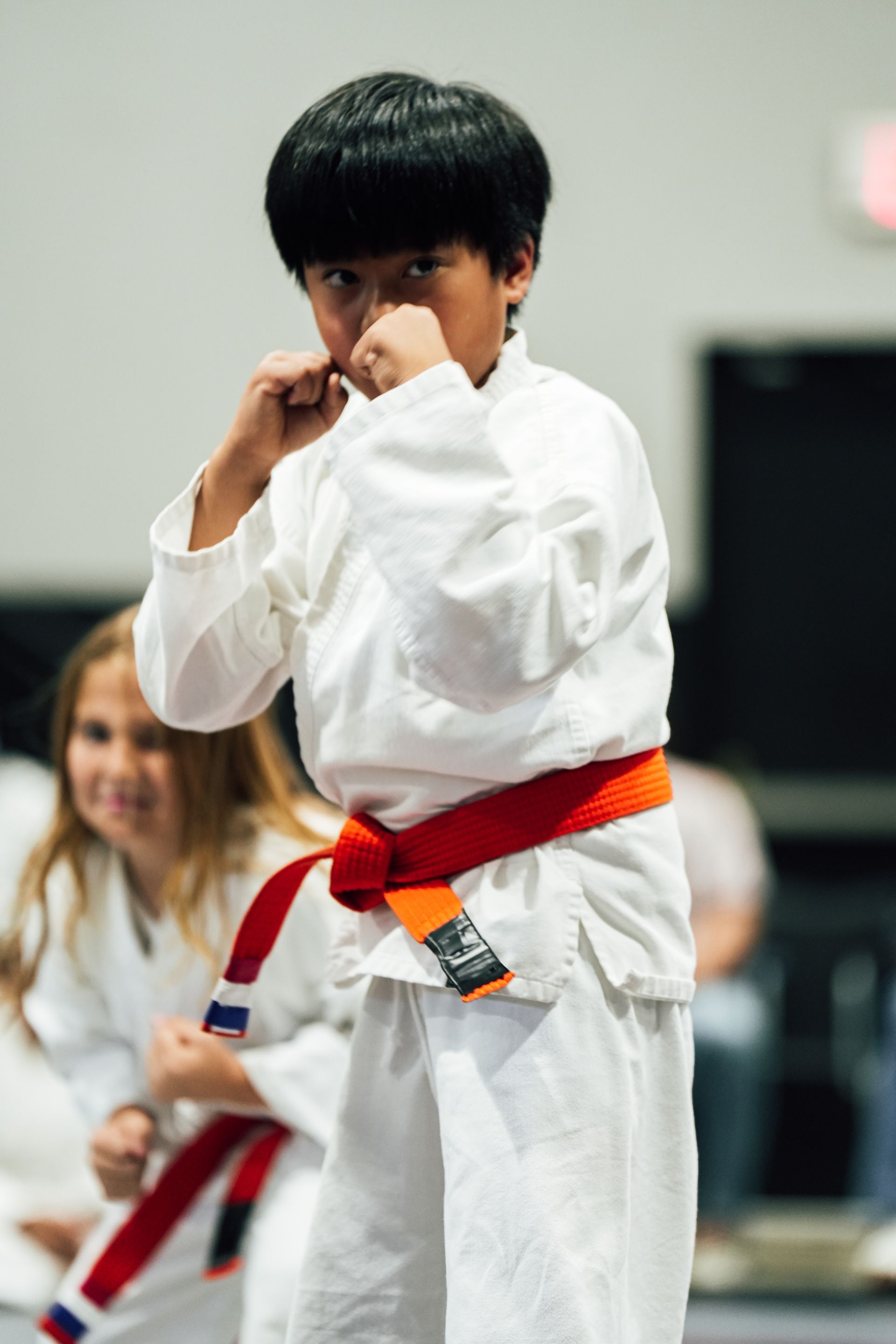 Martial arts class: students in white gis kneeling, instructor in black belt, mirrored room.