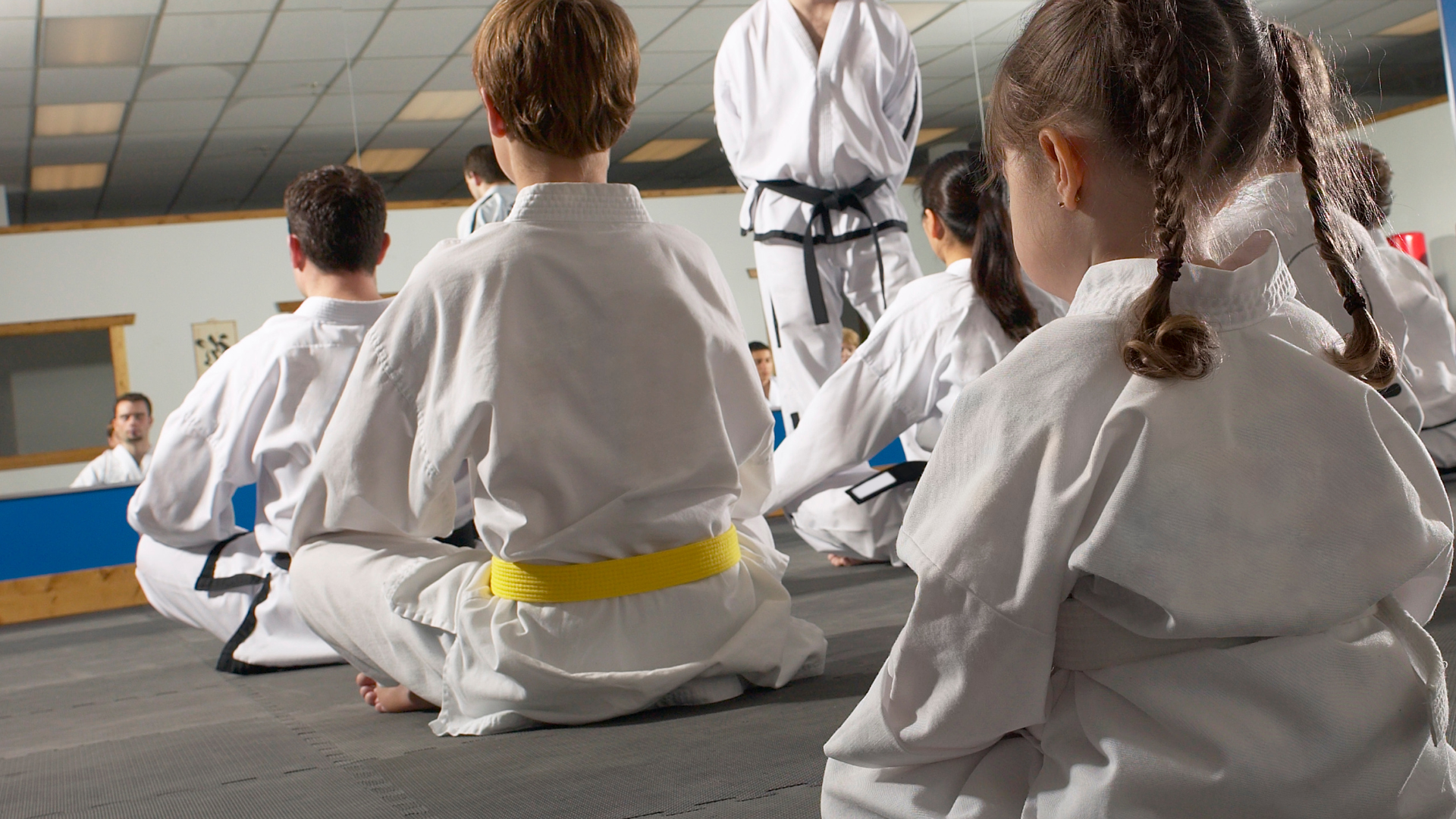Martial arts class: students in white gis kneeling, instructor in black belt, mirrored room.