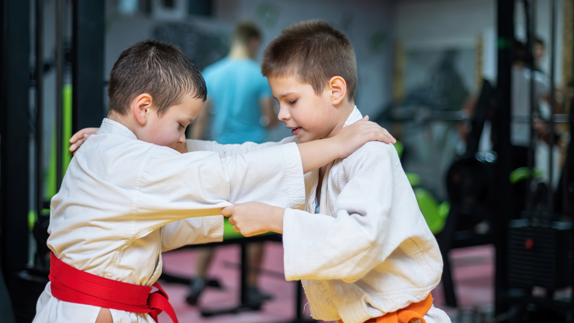 Two boys in karate uniforms practice a move in a gym. One wears an orange belt, the other a red belt.