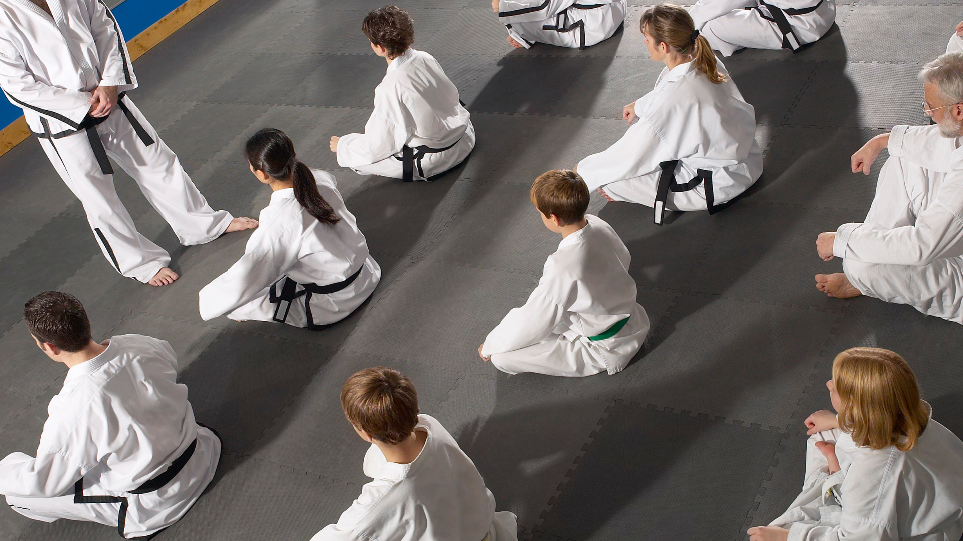 People in white martial arts uniforms sitting on a mat, being addressed by an instructor.
