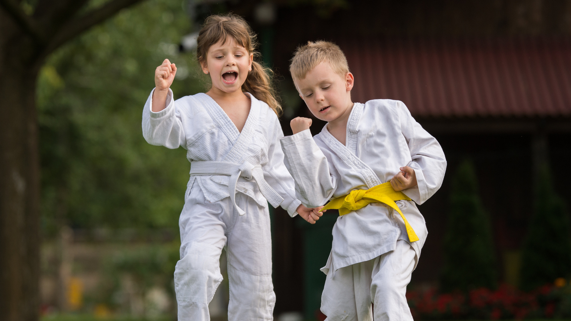 Two children in karate uniforms holding hands, outdoors, one with yellow belt, joyful expressions.