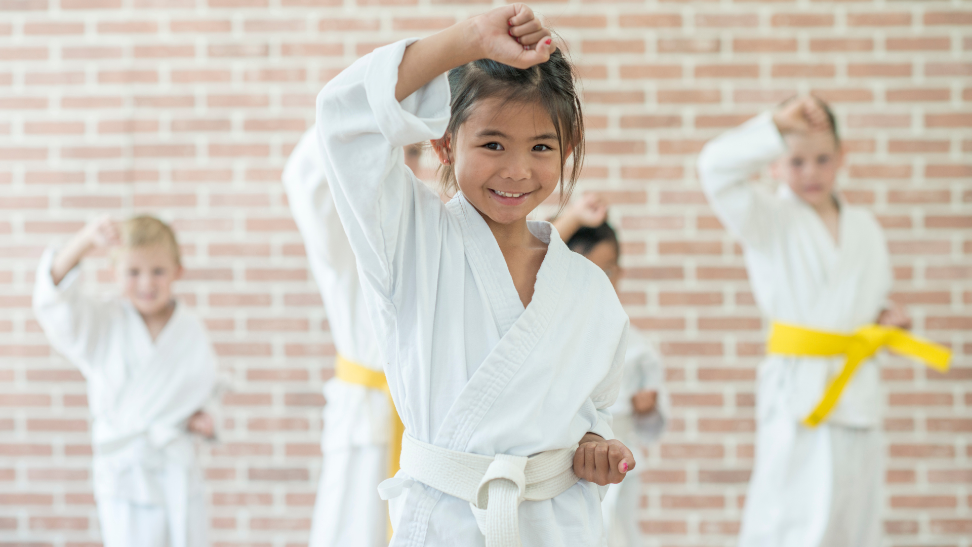 Children in karate uniforms practicing martial arts, smiling. Brick wall background.
