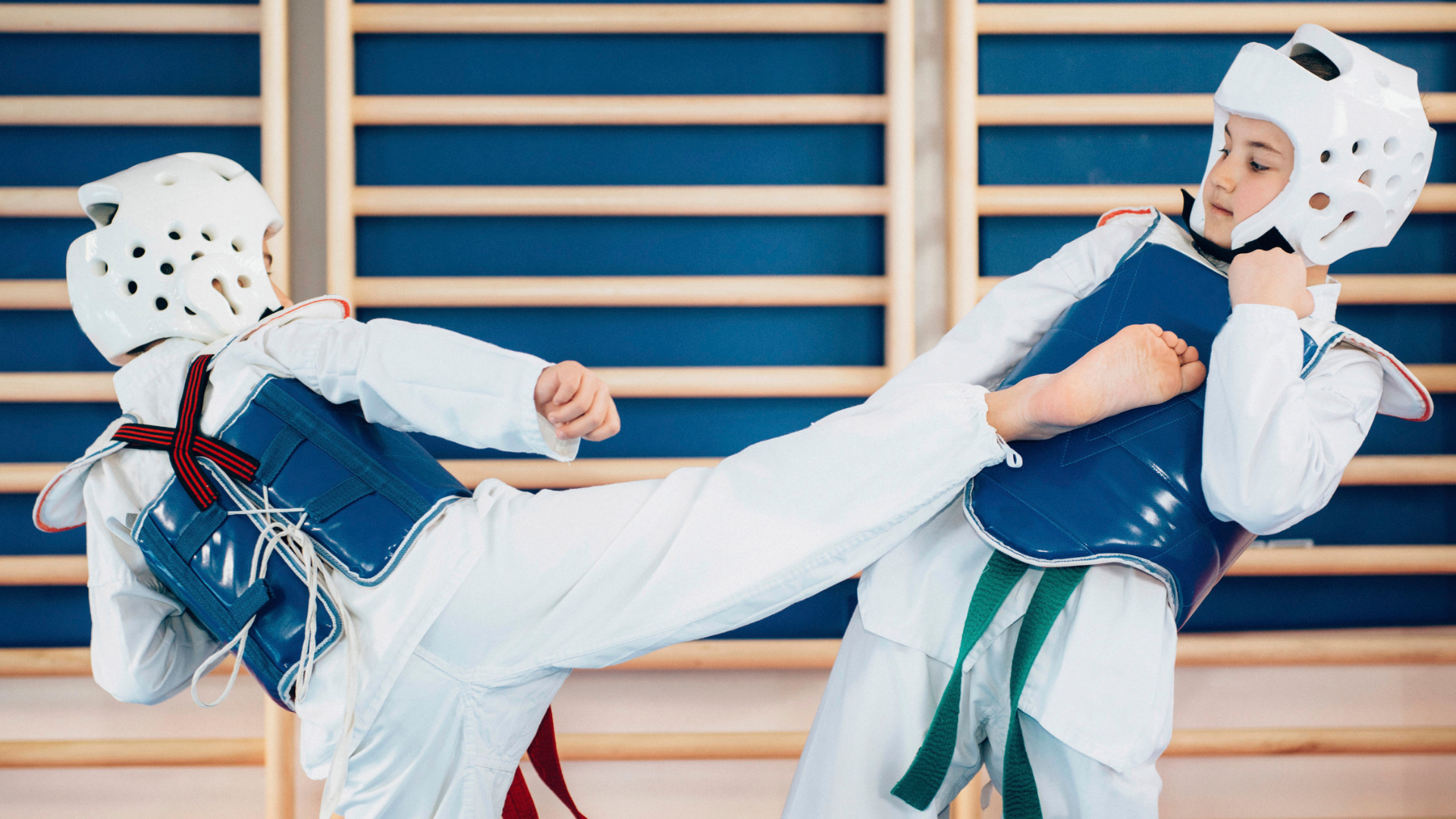 Two people in taekwondo gear sparring in a gym, one kicking towards the other.