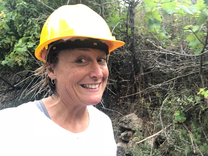 A woman wearing a yellow hard hat is smiling in the woods.