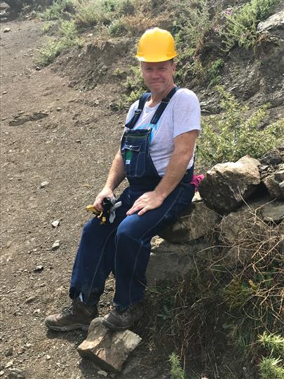 A man wearing overalls and a hard hat is sitting on a rock.