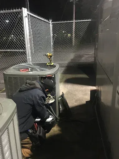 Worker crouches near HVAC equipment by a chain-link fence at night.