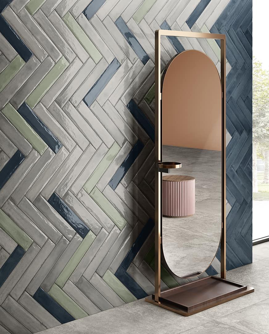 Parete piastrellata a spina di pesce, specchio alto con cornice dorata che riflette uno sgabello rosa e parete color pesca.