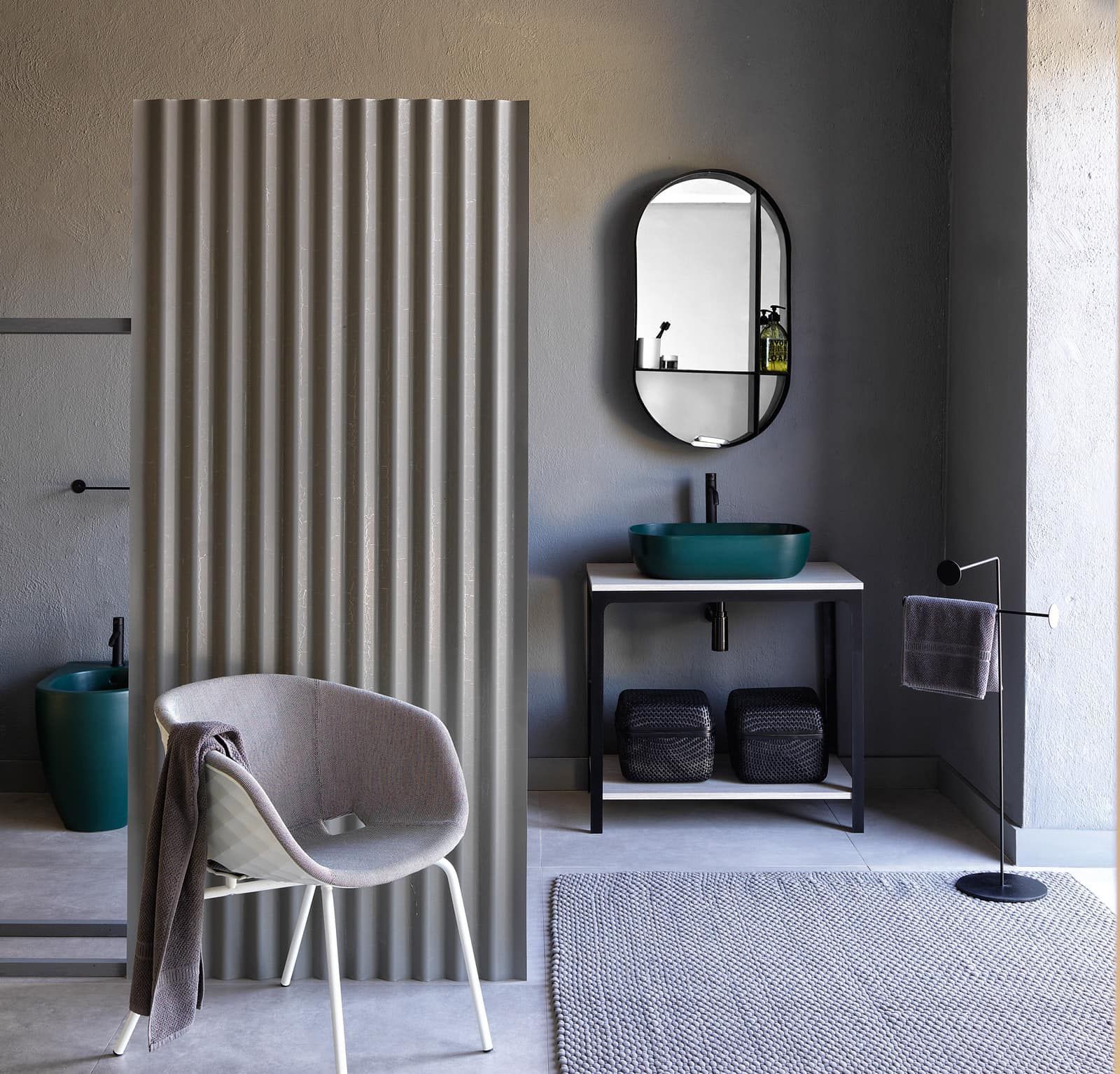 Bagno con pareti grigie, lavandino verde, divisorio ondulato, sedia e specchio ovale.