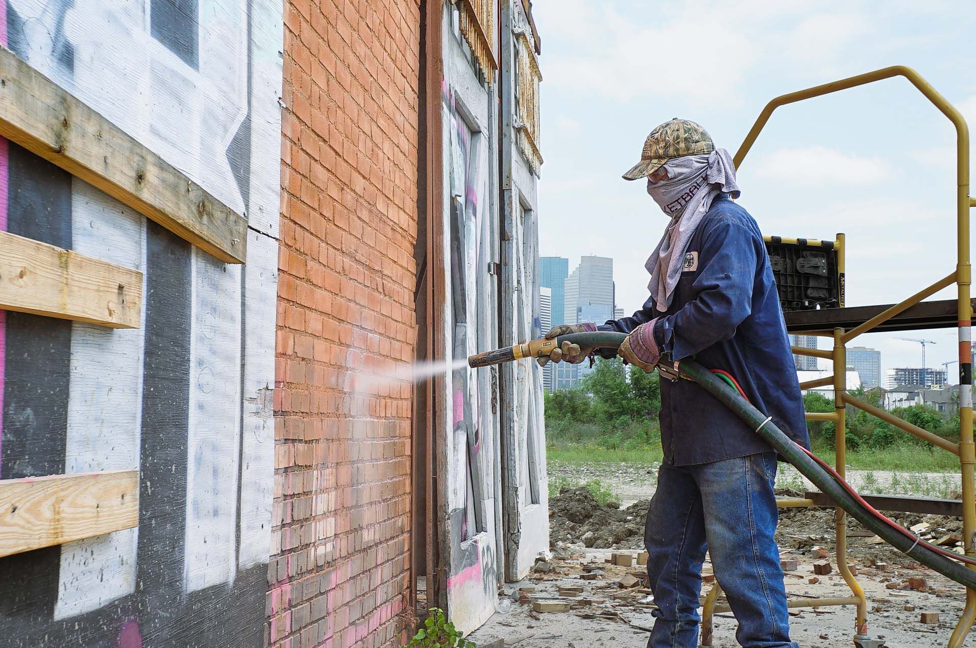 A man is sandblasting a brick wall with graffiti on it.