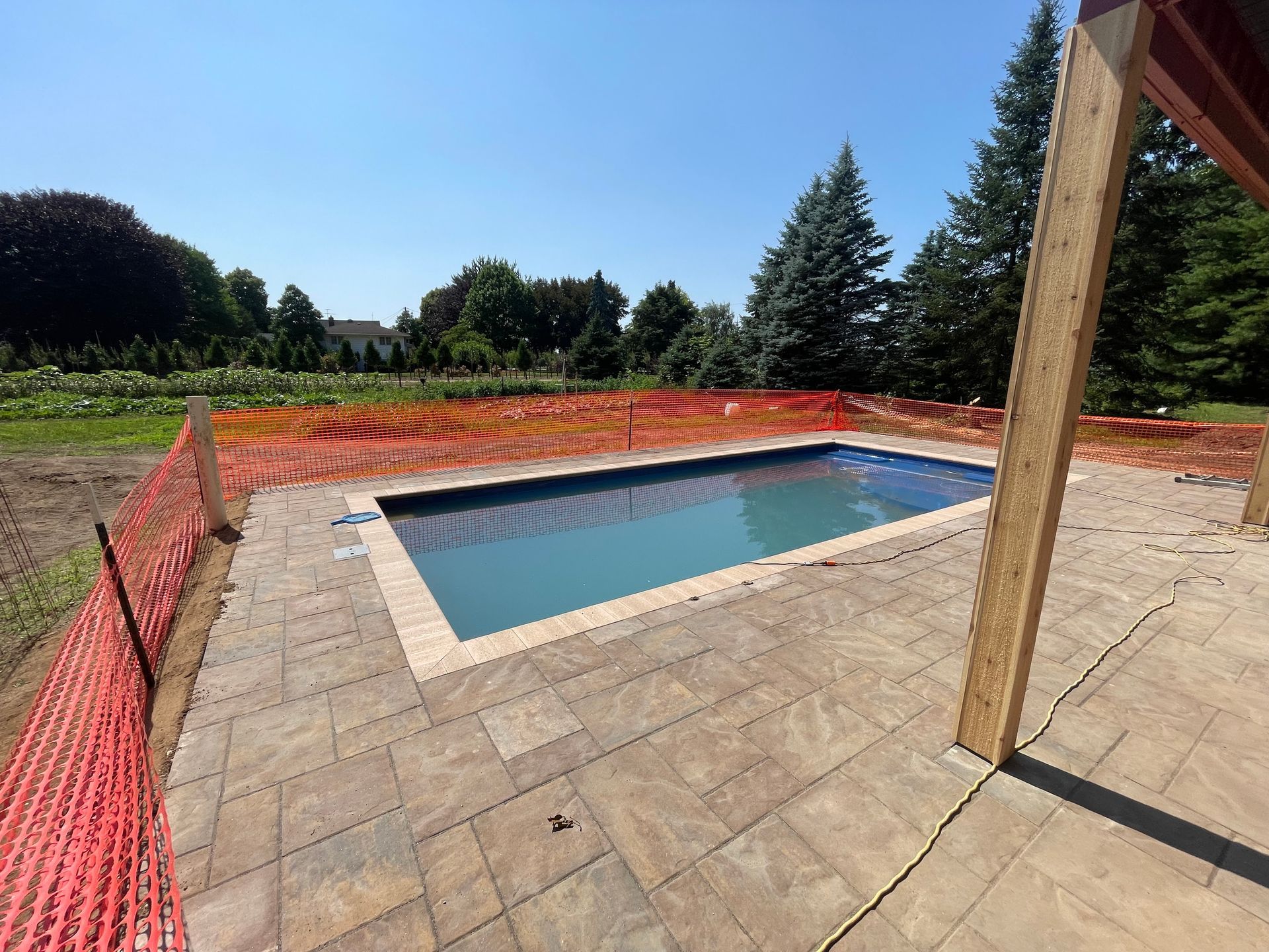 Rectangular pool with blue water and stone patio, orange construction fence, under blue sky.