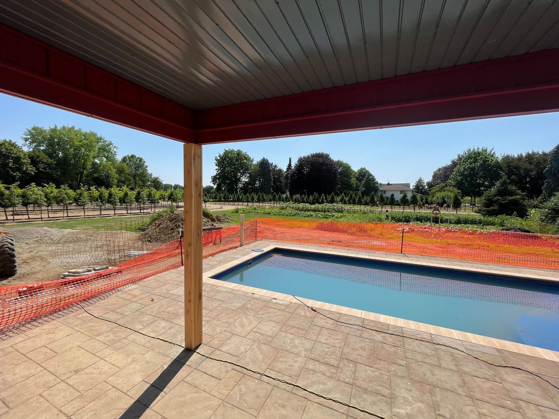 Pool under construction, viewed from a covered patio. Red beams and orange safety fence. Trees and blue sky in background.