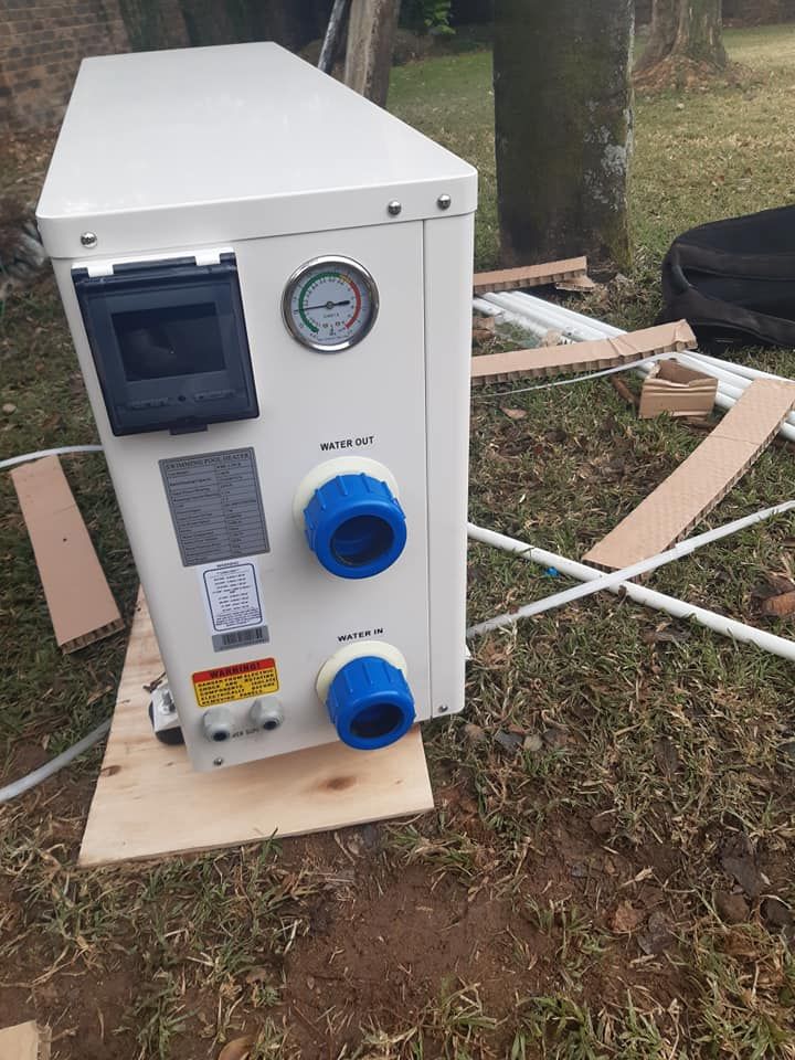 White outdoor heat pump with blue pipes, gauge, and control panel, sitting on wood in grass.