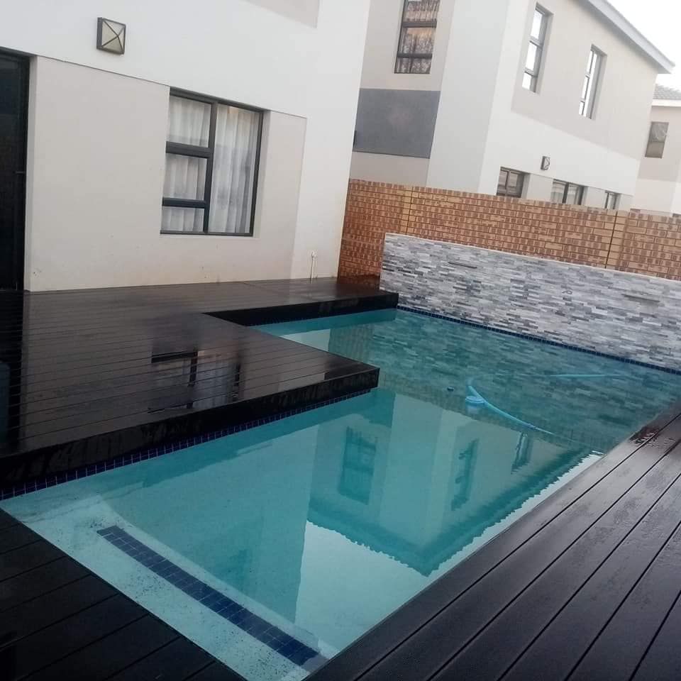 Swimming pool with dark wood decking and a textured stone wall, reflecting a white building.