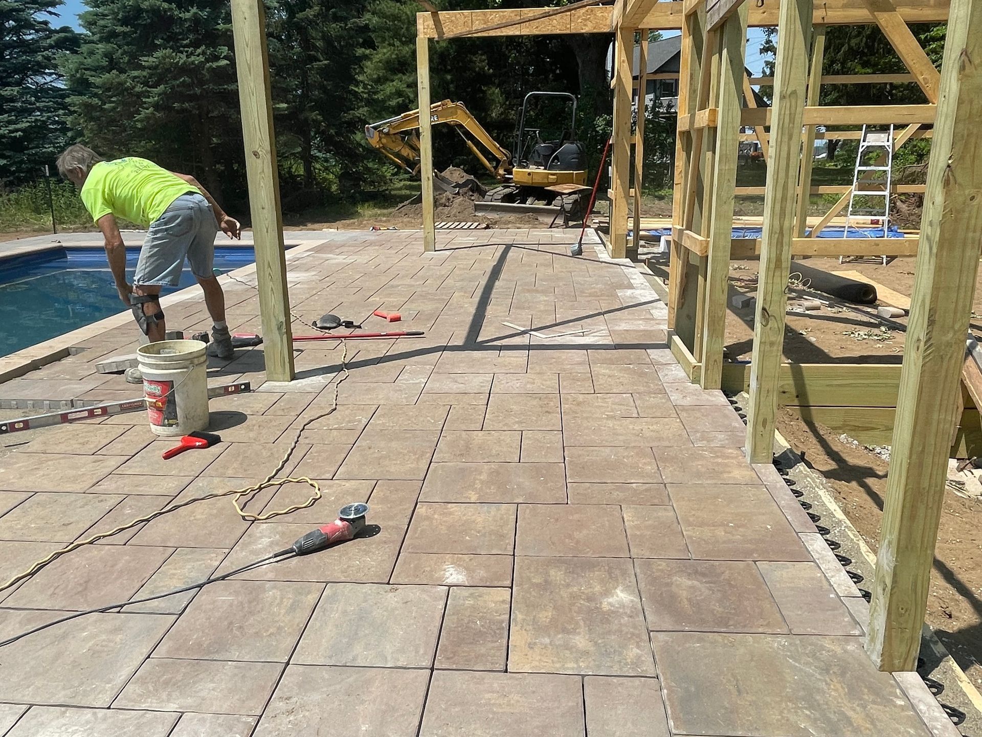 Construction of a patio with pavers; a worker near a pool, wood frame structure in progress.