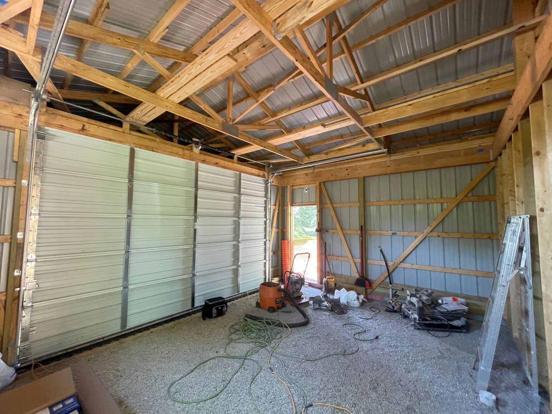 Interior view of a barn under construction with a garage door, tools, and exposed wooden beams.