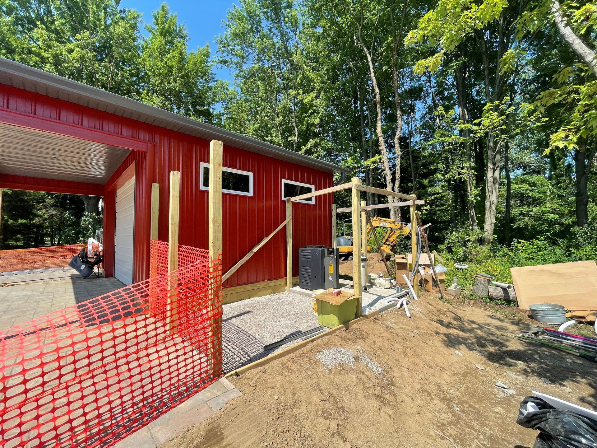 Red metal building under construction with gravel area, safety fencing, and surrounding trees.