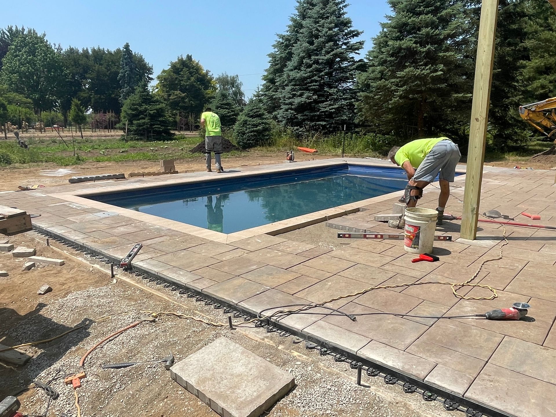 Construction workers install pavers around a rectangular pool. Two workers wear neon vests, one drills. Outdoors, sunny day.