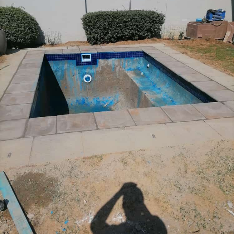 Construction workers installing pavers around a rectangular swimming pool, outdoors on a sunny day.