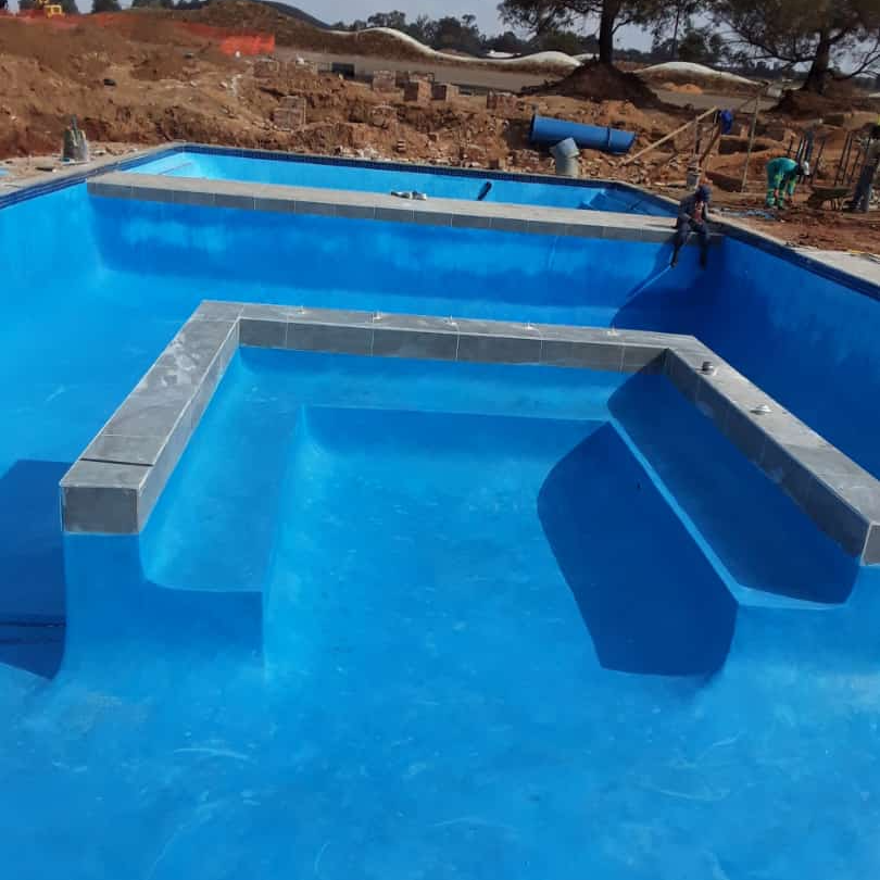 Newly constructed blue swimming pool with steps, concrete borders, and construction site background.