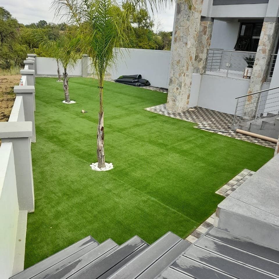 Green artificial turf yard with small palms, next to a building and white fence.