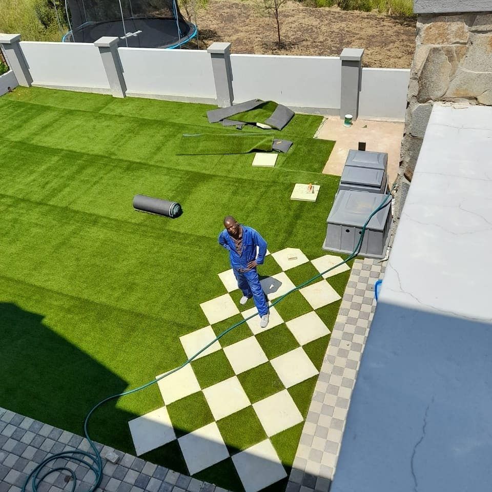 Man installing checkered patio tiles next to green artificial turf in a yard.
