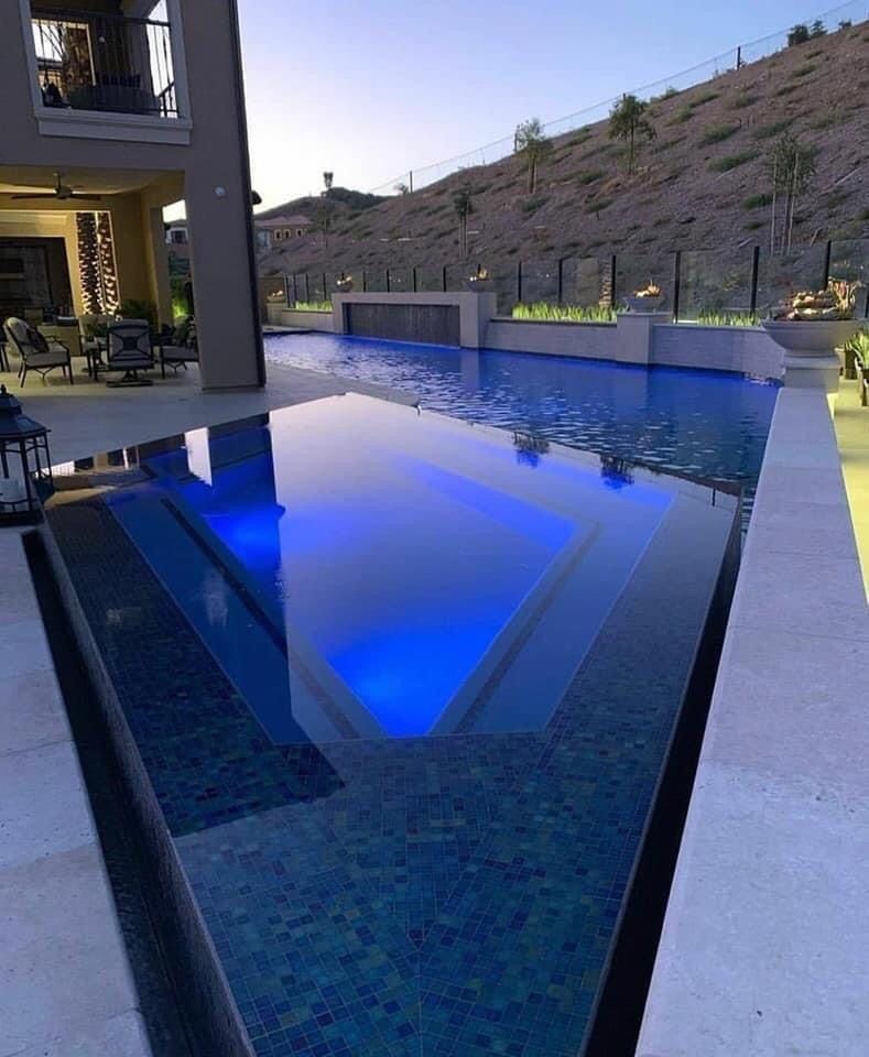 A modern pool with blue underwater lights, surrounded by a patio and a hillside background.